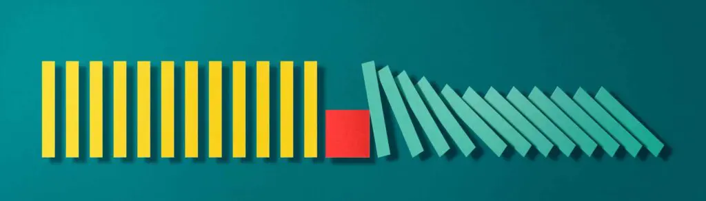 umfallende bunte Dominosteine, in der Mitte durch Quader gestoppt als Symbol für Prävention