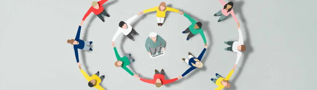 Menschen bilden an den Händen haltend schützende Kreise um eine ältere Frau am Rollator